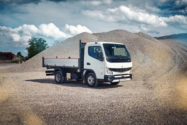 White flatbed truck parked on gravel with piles of dirt in the background.